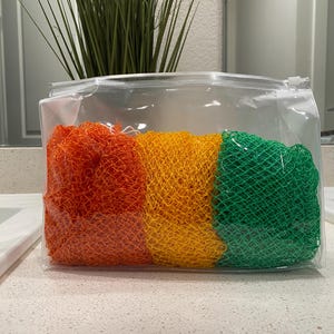 May include: A clear, zippered pouch containing three colorful mesh bath sponges. The sponges are orange, yellow, and green. The pouch is sitting on a white countertop.