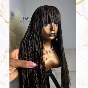 May include: A brown and black braided wig with a blunt bang. The wig is displayed on a mannequin head with a pearl necklace. The text "DSW DAINTY WIG" is visible in the background. The wig is held by a hand with a pink nail polish.