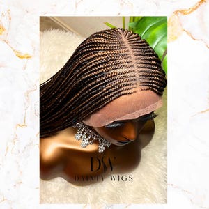 May include: A brown and black braided wig with a lace front is displayed on a mannequin head. The wig features intricate braiding and a natural-looking hairline. The mannequin is adorned with a sparkling necklace, and the brand name "DAINTY WIGS" is visible.