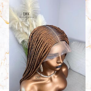 Puede incluir: Una peluca trenzada marrón con una parte delantera de encaje se muestra en una cabeza de maniquí. La peluca presenta una raya en el centro y está peinada con numerosas trenzas pequeñas. El maniquí lleva un collar y pendientes plateados. El texto "DSW DAINTY WIGS" es visible.