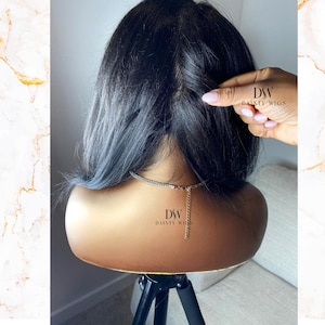 May include: A black wig with blue-tinted ends is displayed on a mannequin head. The wig is styled with a center part. The mannequin is on a black tripod stand. The text "DW DAINTY WIGS" is visible on the mannequin and a hand.