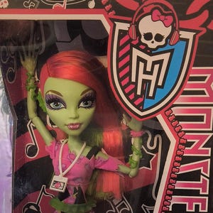 Peut inclure: Poupée Venus McFlytrap de Monster High dans une boîte en plastique transparente. La poupée a la peau verte, les cheveux roux et porte un haut rose et un short noir à motifs. La boîte présente le logo Monster High et le texte "Venus McFlytrap".