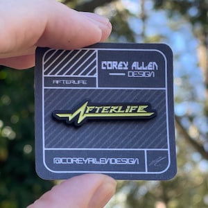 May include: A black and gray pin with the word "AFTERLIFE" in yellow, held by a hand. The pin is on a black and gray card with the text "COREY ALLEN DESIGN" and "@COREYALLENDESIGN".