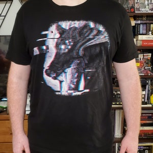 May include: Black t-shirt featuring a stylized wolf design with a 3D glitch effect in white, red, and blue. The graphic is centered on the chest. The shirt has short sleeves and a crew neck.
