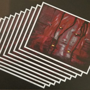 May include: A stack of art prints featuring a dark, surreal illustration. The artwork depicts a face with red hair and glowing yellow eyes, set against a deep red background. The prints have white borders and are stacked on a black surface.