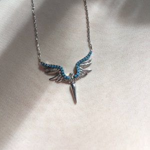 May include: Silver necklace with a pendant featuring angel wings with turquoise stones and a pointed tip.