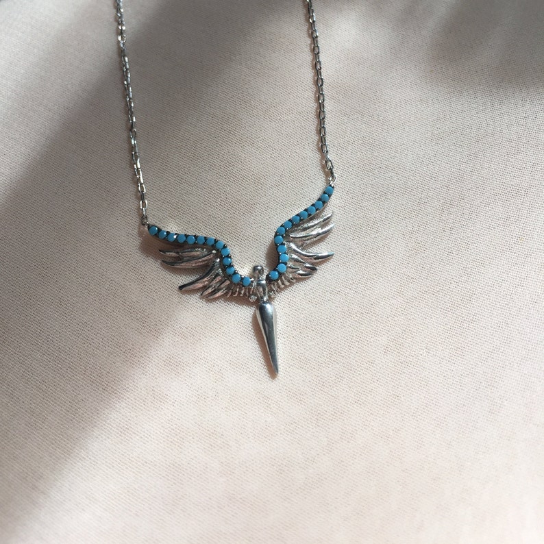 May include: Silver necklace with a pendant featuring angel wings with turquoise stones and a pointed tip.