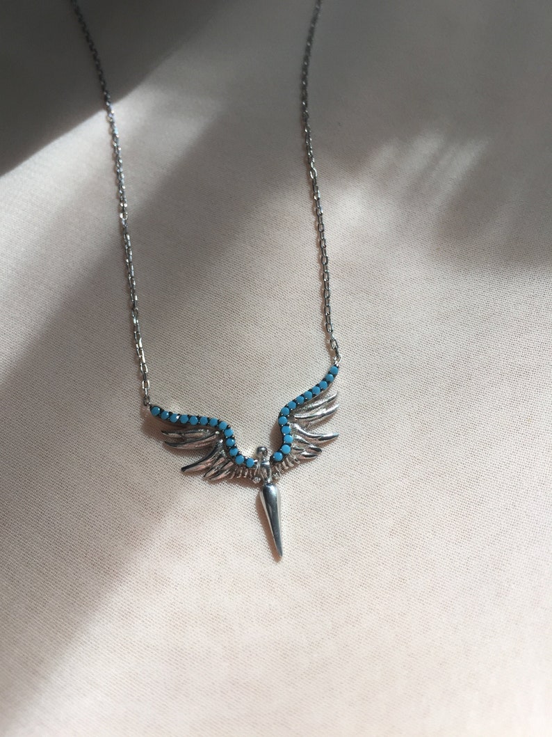 May include: A silver necklace with a turquoise stone pendant shaped like a pair of wings with a pointed tip.