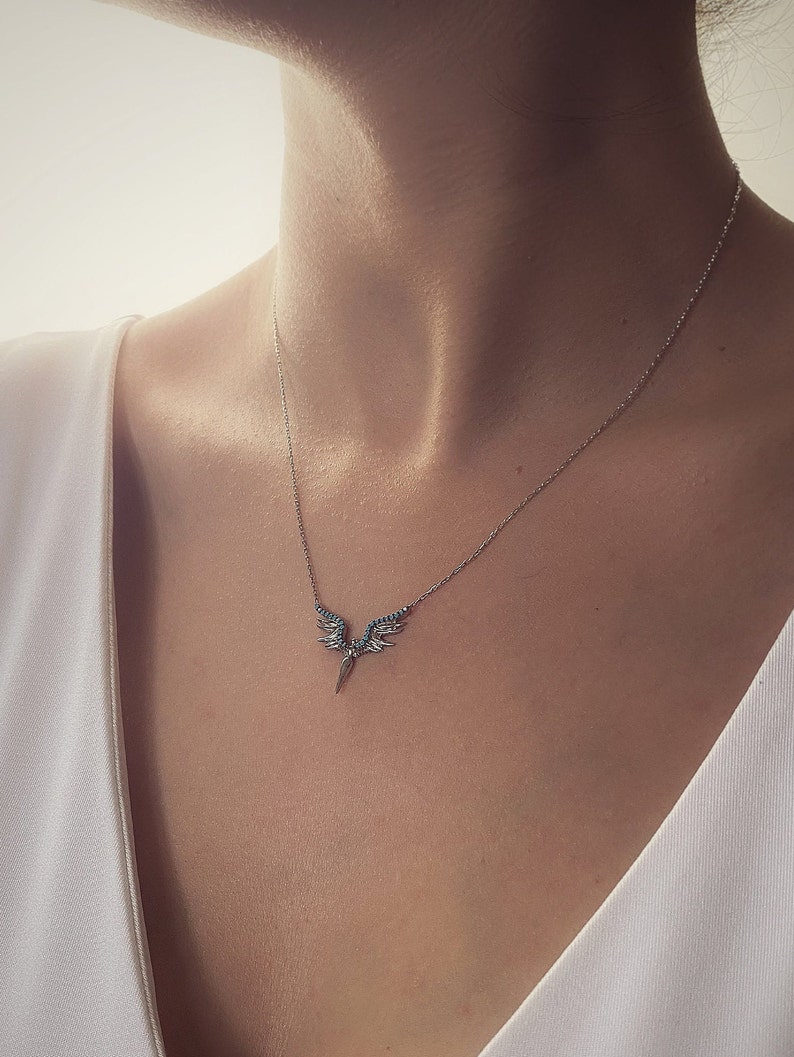 May include: A silver necklace with a delicate winged pendant featuring blue gemstones. The pendant is shaped like a pair of wings with a pointed tip.