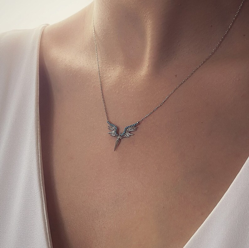 May include: A silver necklace with a pendant featuring a pair of angel wings with turquoise stones and a pointed tip.