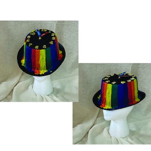 May include: A black top hat with a rainbow-colored, dripped paint design. The hat features gold-colored numbers and clock hands. The hat is displayed on a white mannequin head.