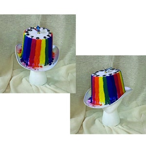 May include: A whimsical, colorful hat with a rainbow design. The hat features a white brim and a cylindrical crown painted with vertical stripes of red, orange, yellow, green, blue, and purple. A clock is on top of the hat.