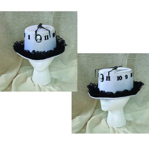 May include: A white top hat with a clock face design. The hat features black numbers, clock hands, and a decorative black lace trim around the brim. The hat is displayed on a white mannequin head.