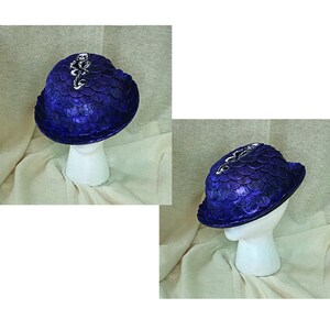 May include: A dark blue hat with a scale-like texture, topped with a silver emblem featuring a skull and snake design. The hat is displayed on a white mannequin head, showcasing its shape and details.