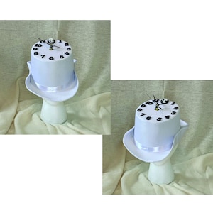 May include: A white top hat with a clock face design. The hat features a white brim, a white ribbon, and a cylindrical crown with black numbers. The clock hands are black and the hat is displayed on a white mannequin head.