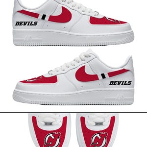 May include: White low-top sneakers featuring red accents, a red Nike swoosh, and the word DEVILS in black lettering. The toe box displays a red panel with the New Jersey Devils logo. The design includes white laces and a white rubber sole.