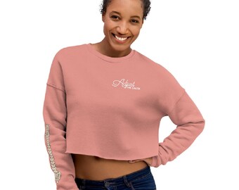 Op de afbeelding: Een cropped, stoffig roze sweatshirt met de tekst "Adjust the Cause" in sierlijke letters op de borst. De linkermouw heeft een ruggengraatontwerp. Het model draagt een blauwe jeans.