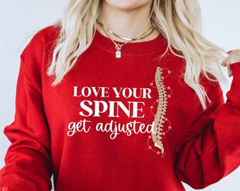 Pode incluir: Camisola vermelha com um gráfico branco que diz "Love your spine get adjusted" e uma imagem de desenho animado de uma coluna vertebral com corações.