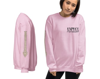 Op de afbeelding: Roze sweatshirt met de tekst "EXPECT miracles" op de voorkant en een wervelkolom afbeelding op de mouw.