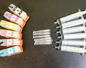 May include: An assortment of printer ink cartridges in black, cyan, magenta, yellow, gray, and red, alongside several syringes and needles. The ink cartridges have orange clips and color labels. The syringes are white with black measurement markings.