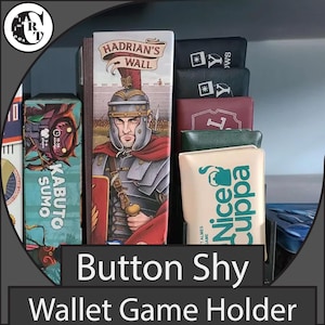 May include: A display of board game boxes and wallets. The "Hadrian's Wall" box features a Roman soldier illustration. Other boxes include "Kabuto Sumo" and wallets with "Nice Cuppa" and "Y" logos. The text "Button Shy Wallet Game Holder" is also visible.