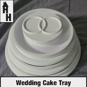 May include: A white circular tiered wedding cake tray featuring a raised interlocking ring design on the top surface. The tray has multiple concentric levels with smooth edges and a solid white finish. The logo AH is visible in the corner.