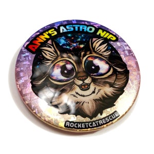 Può includere: Un bottone rotondo con finitura olografica presenta un gatto dei cartoni animati con una galassia negli occhi. Il testo "Ann's Astro Nip Rocketcatrescue" è stampato intorno al gatto.