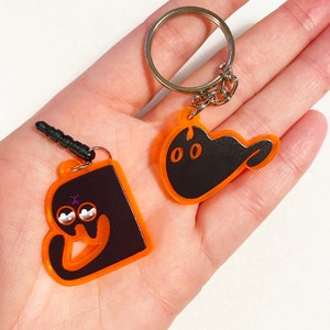 Puede incluir: Dos llaveros con forma de gato negro con contornos naranjas. Un llavero tiene un anillo para llaves y el otro tiene un dije de teléfono negro y morado.
