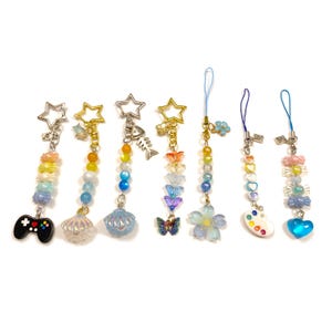 Op de afbeelding: Een verzameling kleurrijke bedelarmbanden met diverse hangers, waaronder een ster, een zeeschelp, een vlinder en een gamecontroller. De bedels hebben een mix van pastelkleurige en levendige kralen, met zilver- en goudkleurige hardware.
