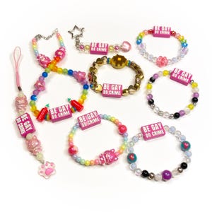 May include: A collection of colorful beaded bracelets and charms. The bracelets feature rainbow, pink, and gold beads, with charms including stars, hearts, and the text "BE GAY DO CRIME". A pink phone charm with a paw print is also visible.