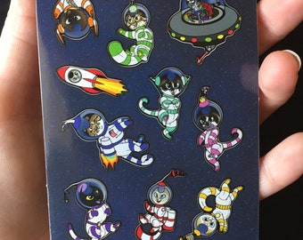 Puede incluir: Una hoja de pegatinas con gatos de dibujos animados vestidos de astronautas y un cohete, sobre un fondo azul oscuro estrellado. Los gatos están en diferentes poses y diseños de trajes, con colores como naranja, verde, rosa y morado.