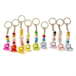 Könnte beinhalten: Eine Sammlung farbenfroher Schlüsselanhänger mit einem Glückskatzen-Design. Jeder Schlüsselanhänger hat einen Metallring, eine Perlenkette und eine kleine, durchscheinende Katzenfigur in verschiedenen Farben, darunter Rosa, Rot, Gelb, Grün, Blau und Lila.