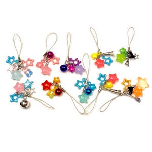 Puede incluir: Una colección de colgantes en forma de estrella de colores con lazos de metal plateado. Los colgantes presentan varios colores, incluyendo rosa, azul, verde y naranja, y algunos tienen pequeñas campanas o cuentas. Adecuado para la fabricación de joyas o como accesorios decorativos.