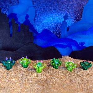 May include: Seven small green cactus figurines with painted faces and purple flowers on top. The figurines are arranged in a row on a sandy surface. The background is a blue and black landscape.