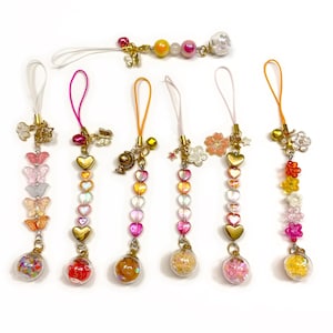 May include: A selection of decorative phone charms in various colours. Each charm features a clear glass orb filled with decorative elements, paired with beads, hearts, butterflies, and floral accents. The charms are in shades of pink, yellow, orange, and gold.