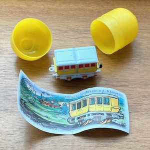 May include: A yellow toy train car with a white roof and red accents, next to a yellow plastic capsule and a paper with a train illustration. The train car is on a wooden surface.