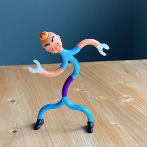 May include: A bendable action figure with a bald head, wearing a blue shirt and pants, with orange arms and white hands. The figure has a purple midsection and black shoes. It is posed on a wooden surface.
