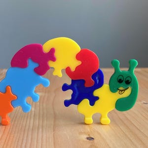 May include: A colorful caterpillar puzzle toy. The puzzle is made of interlocking pieces in various colors, including orange, blue, pink, yellow, red, and green. The caterpillar has a smiling face and is positioned on a wooden surface.