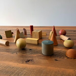 May include: A collection of various wooden geometric shapes including spheres, cones, cylinders, cubes, and rectangular prisms in natural wood tones, muted greens, and reddish-brown hues scattered across a dark brown wooden surface.
