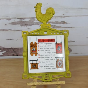 May include: A yellow cast iron rooster sign with a tile insert listing the rules of a tavern. The tile has a white background with black text and illustrations of signs for the Half Moon Tavern, the Drum Tavern, and the Tile Tavern. The text reads: "RULES of the TAVERN 4 pence a night for bed 6 pence with potluck 2 pence for horse keeping No more than five to sleep in one bed No boots to be worn in bed No razor grinders or tinkers taken in No dogs allowed in the kitchen Organ grinders to sleep in the wash house"