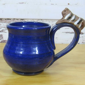 May include: A blue ceramic mug with a brown and white striped animal figurine on the handle. The mug has a rounded body and a wide rim.