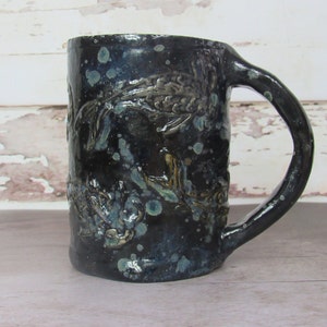 May include: A black ceramic mug with a blue and green glaze. The mug has a handle and is decorated with a raised design of fish.