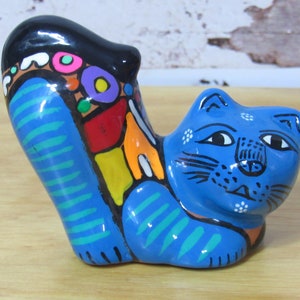 May include: A blue ceramic cat figurine with colorful stripes and geometric patterns on its back. The cat is lying down with its head turned to the side and its eyes closed.