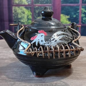 May include: Black ceramic teapot with a rooster design. The teapot has a lid and a metal handle. The rooster is depicted in white and gray with a red comb. The teapot has a metal coil around the body and sits on three small feet.