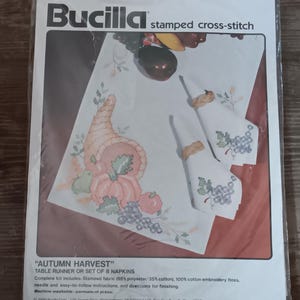 May include: A Bucilla stamped cross-stitch kit for an "Autumn Harvest" table runner and eight napkins. The design features a cornucopia, pumpkins, and grapes. The kit includes stamped fabric, embroidery floss, needle, and instructions.
