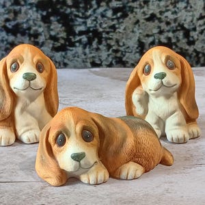 May include: Three small ceramic dog figurines with tan and white fur, long floppy ears, and large dark eyes. Two of the dogs are in a sitting position, while one dog is lying down in the front center. Each figurine has a green nose and paws.