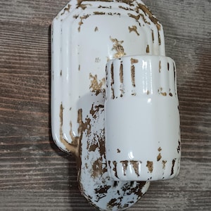 May include: White ceramic wall sconce with a distressed, aged appearance. The sconce features a rectangular backplate and a cylindrical cup holder, both with gold-colored accents. The design suggests a vintage or antique aesthetic.