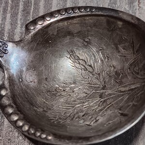 May include: Close-up of an antique silver serving spoon. The spoon's bowl features an engraved wheat and floral design. The rim is decorated with small, raised beads. The handle has an ornate, scrolled design.