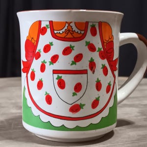 May include: White ceramic mug with a whimsical design featuring a strawberry-adorned apron. The mug has a red rim, a green base, and a brown handle. The apron design includes red strawberries and orange accents.