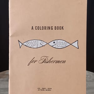 May include: A coloring book for fishermen. The cover is tan with black text and a graphic of two fish. The text reads "A Coloring Book for Fishermen." The book is propped up on a wooden easel.